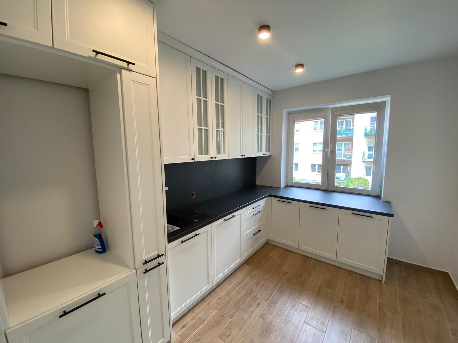 Biała kuchnia na wymiar z czarnym blatem i płytkami, widok na okno z zabudową, drewnopodobna podłoga i minimalistyczne uchwyty szafek.