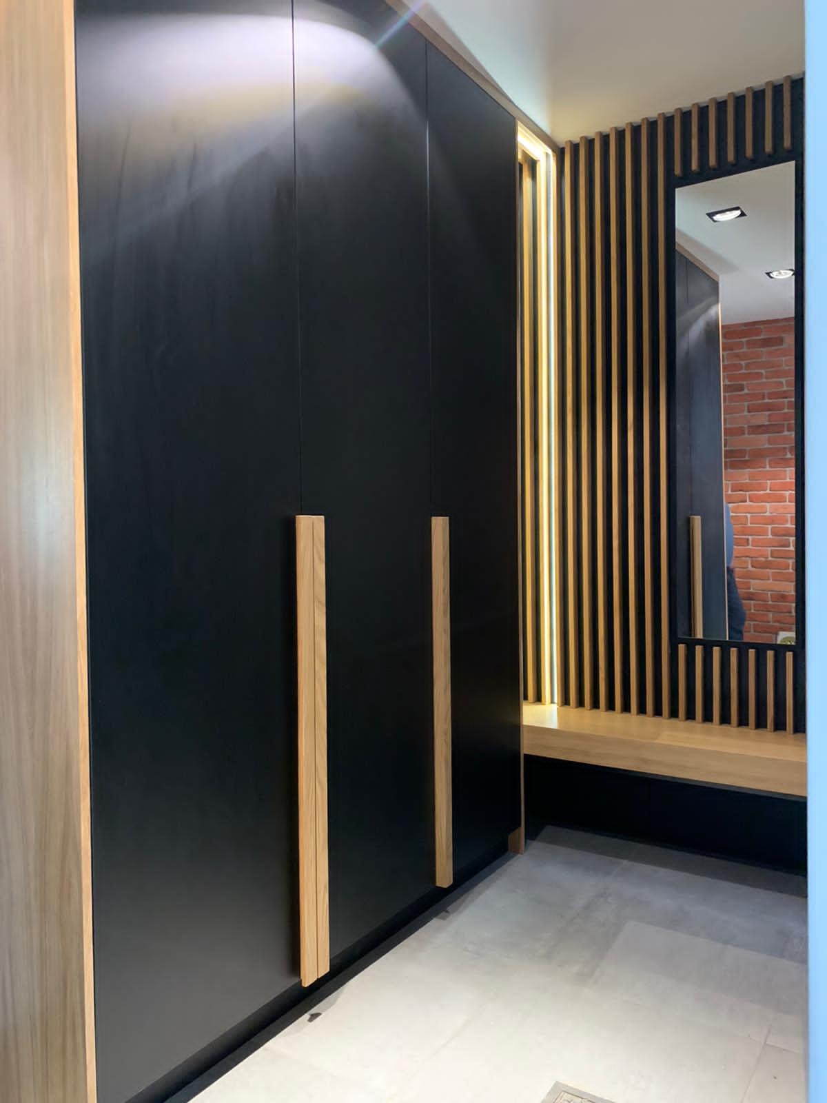 Czarna szafa wnękowa z drewnianymi uchwytami, oświetleniem LED i lustrem w przedpokoju z ceglaną ścianą. Minimalistyczny design, jasna podłoga.