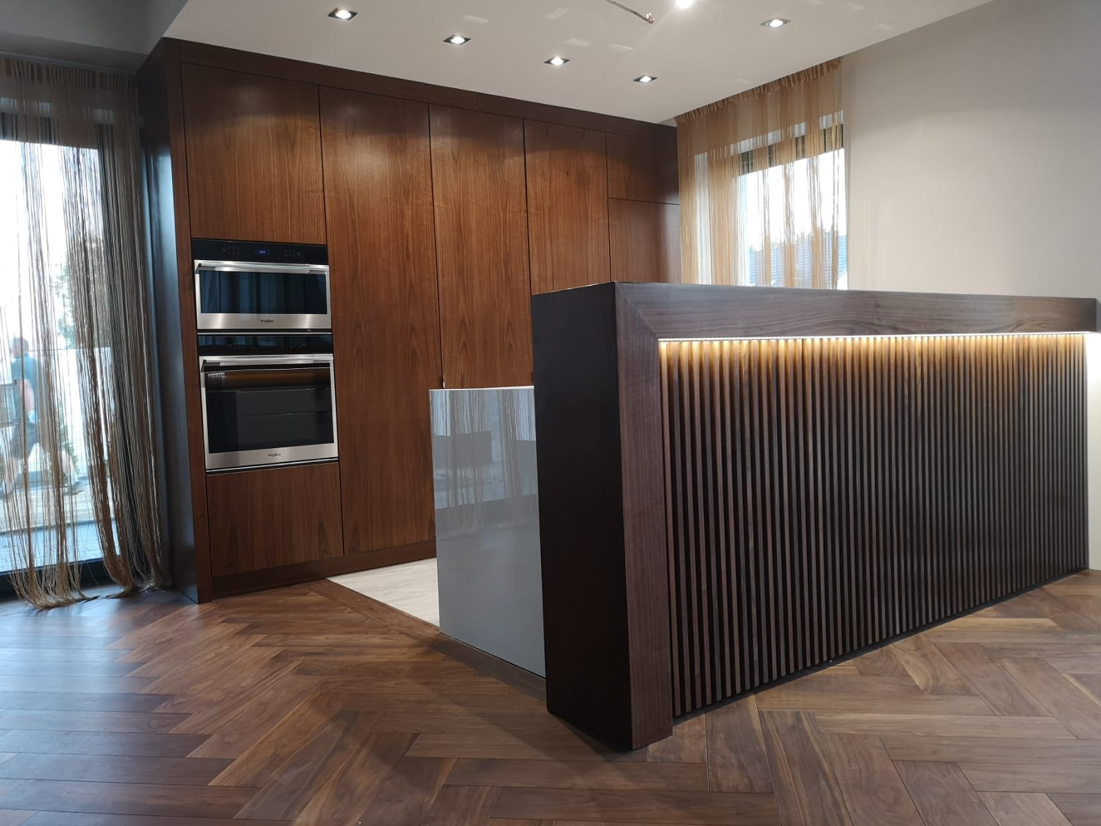 Nowoczesna kuchnia z drewnianymi frontami, zabudowaną lodówką i barkiem z oświetleniem LED na tle parkietu ułożonego w jodełkę.