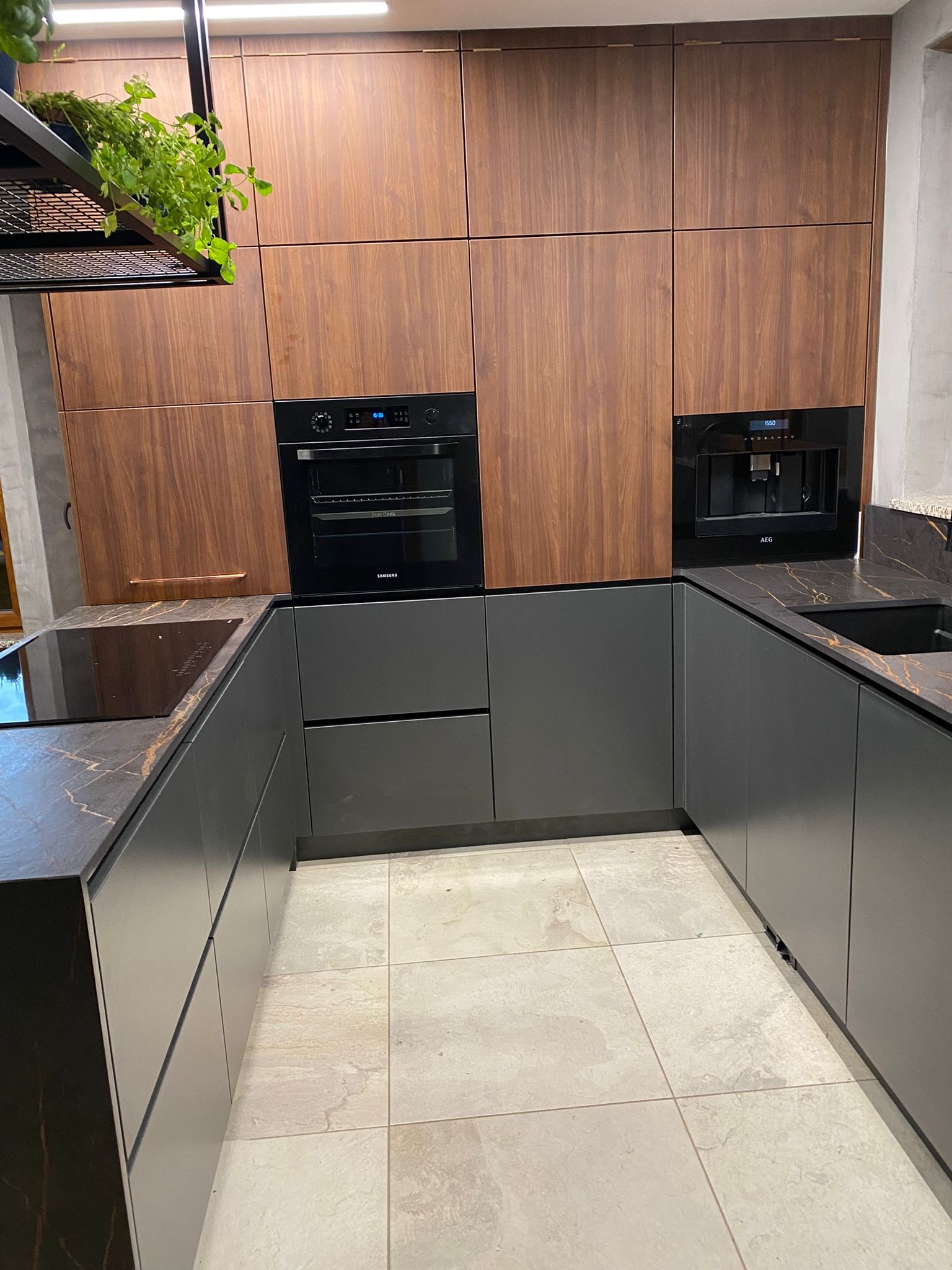 Nowoczesna kuchnia w odcieniach szarości i drewna z wbudowanym sprzętem AGD. Blat z kamienia z wyraźnym żyłkowaniem. Roślina w metalowej konstrukcji nad blatem.