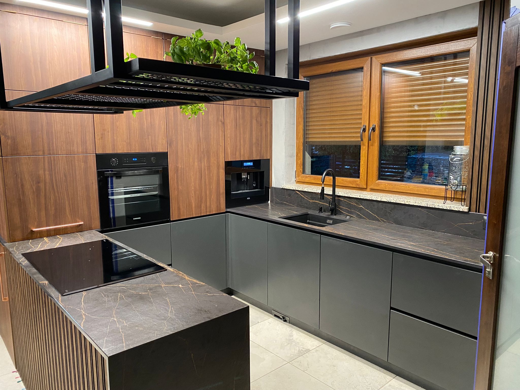 Nowoczesna kuchnia z drewnianymi szafkami górnymi, szarymi dolnymi i blatem z efektem marmuru. Nad wyspą zawieszona czarna konstrukcja z roślinami.