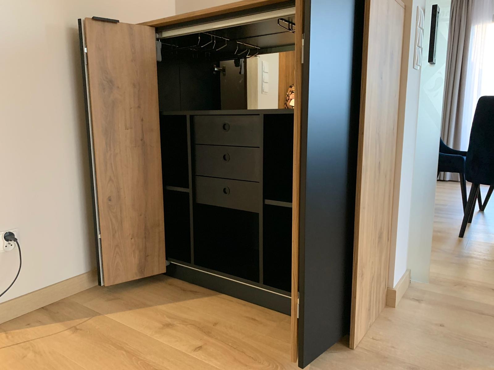 Otwarta szafa wnękowa z drewnianymi drzwiami i czarnym wnętrzem, w którym widoczne są półki, szuflady i wieszaki na kieliszki. Szafa stoi na drewnianej podłodze.