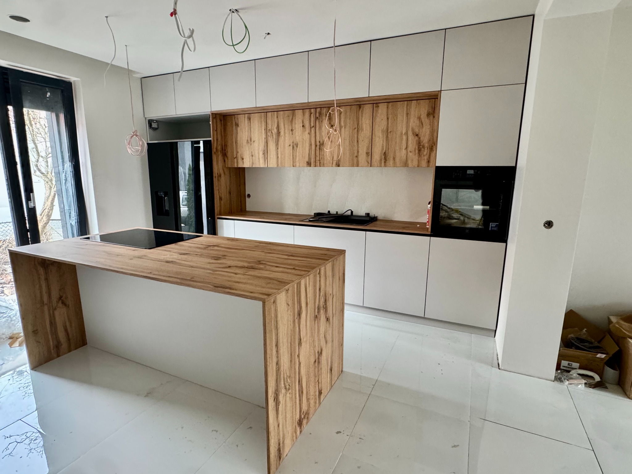 Nowoczesna kuchnia z wyspą: białe szafki górne i dolne, drewniane fronty wyspy i szafek wiszących, czarny sprzęt AGD. Widoczne kable zwisające z sufitu.