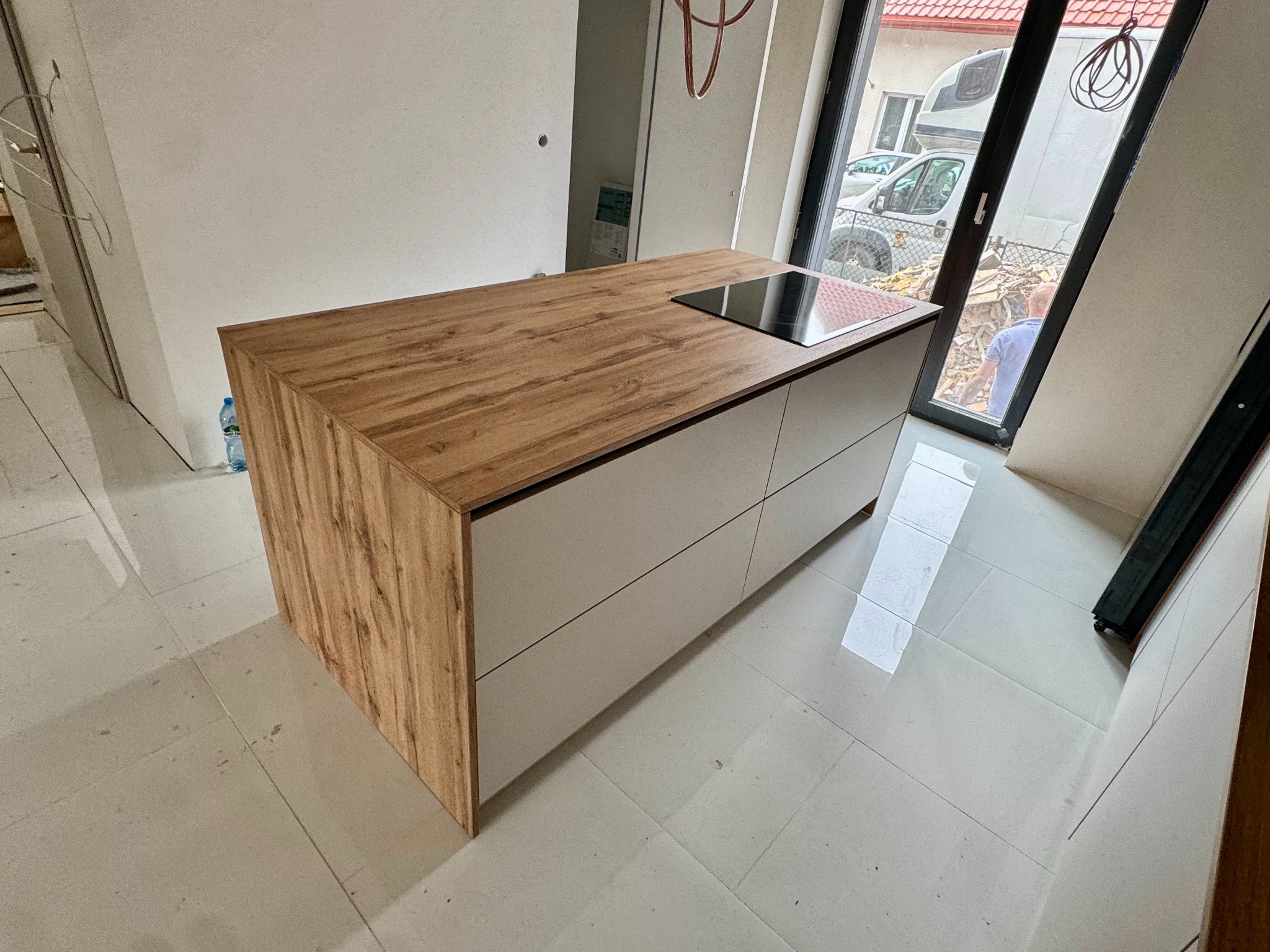 Wyspa kuchenna z drewnianym blatem i płytą indukcyjną w nowym wnętrzu. Białe fronty szuflad, jasna podłoga. Widok na zewnątrz przez okno.