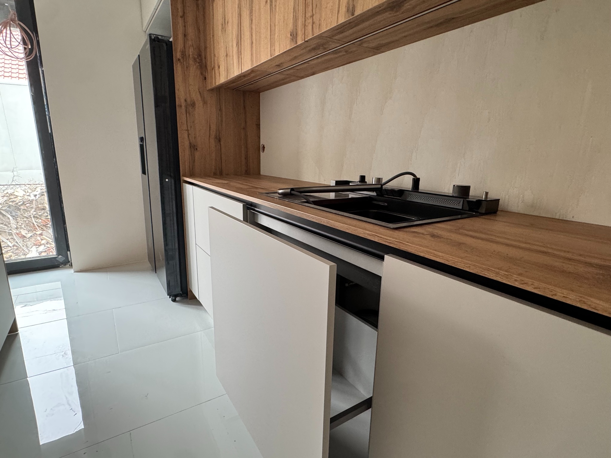 Nowoczesna kuchnia z drewnianymi szafkami i białymi frontami. Widoczny zlew i fragment lodówki. Minimalistyczny design, jasne wnętrze w trakcie montażu.