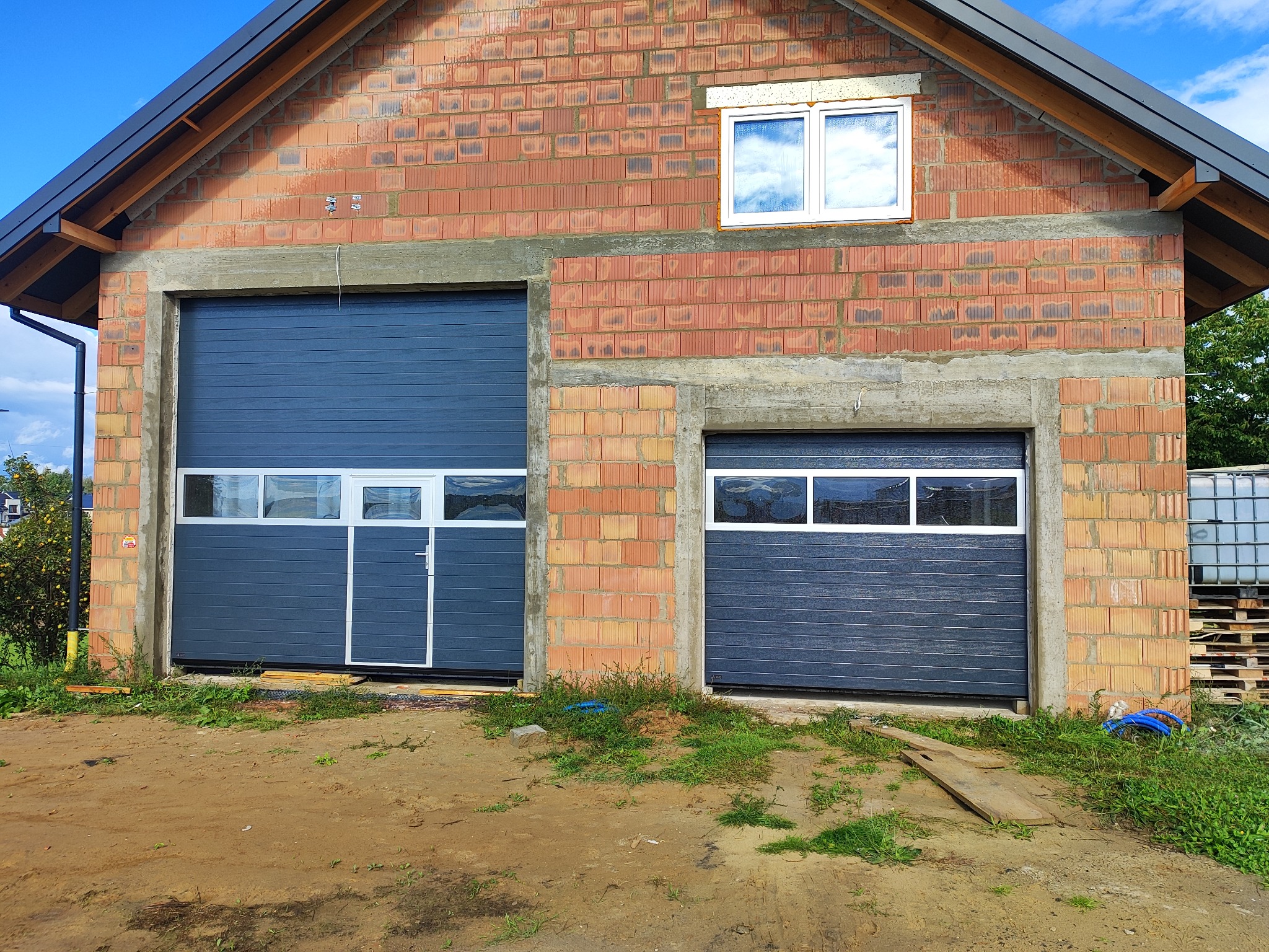 Dwie szare bramy garażowe segmentowe z oknami w surowym budynku z czerwonej cegły. Jedna brama z drzwiami. Widoczne palety i zbiorniki na wodę w tle.