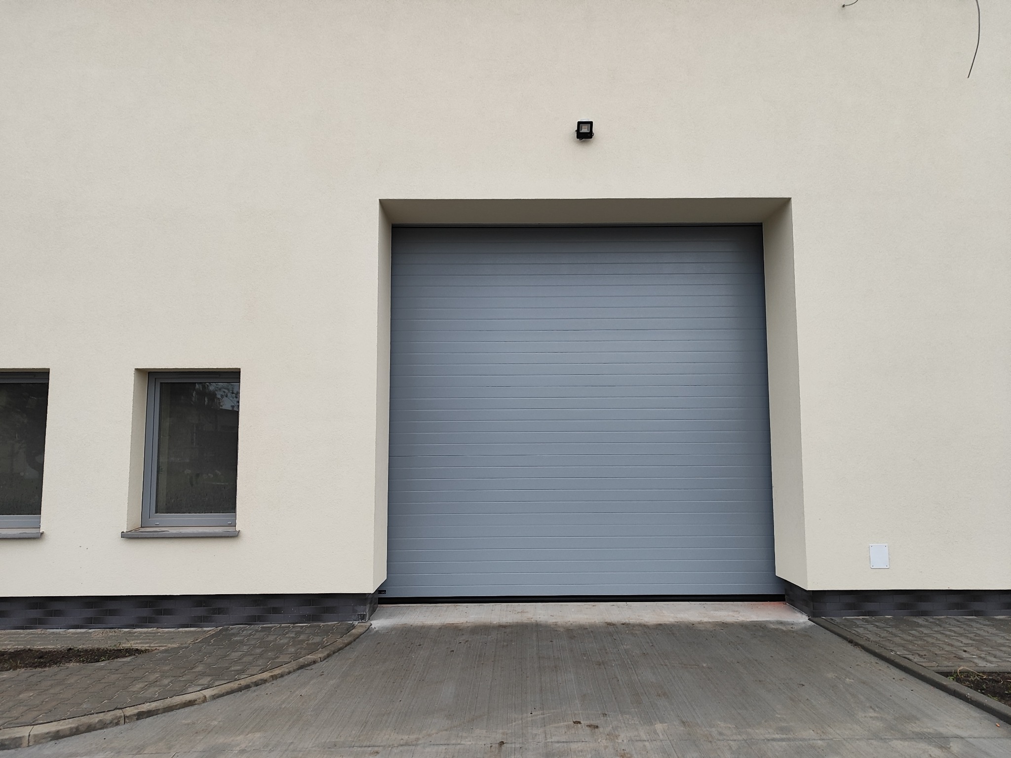 Szara brama garażowa segmentowa w jasnej elewacji budynku z dwoma oknami po lewej stronie. Widoczny fragment podjazdu i lampa nad bramą.