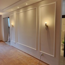 PrimeFix Homes OLEKSANDR DIACHUK - Elegancko wykończone wnętrze z panelami ściennymi, oświetleniem punktowym i drewnianą podłogą w jodełkę. Widoczne gniazdka elektryczne i fragment łazienki w tle.