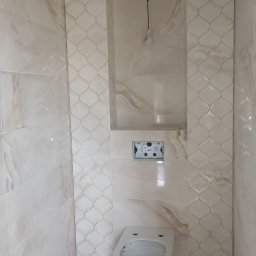 PrimeFix Homes OLEKSANDR DIACHUK - Wykończona łazienka z beżowymi płytkami imitującymi marmur, wisząca toaleta i wnęka w ścianie. Widoczne elementy instalacji podtynkowych i dekoracyjne pasy płytek.