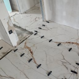 PrimeFix Homes OLEKSANDR DIACHUK - Układanie płytek podłogowych z marmurowym wzorem, z użyciem klipsów poziomujących. W tle ściany w trakcie szpachlowania i narzędzia glazurnicze.