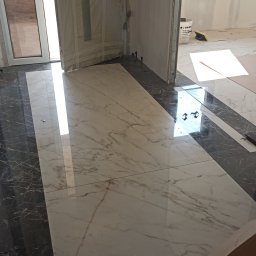 PrimeFix Homes OLEKSANDR DIACHUK - Położona, błyszcząca podłoga z marmurowych płytek w odcieniach bieli i szarości, kontrastująca z czarnymi płytkami przy ścianach. Widoczne drzwi w trakcie montażu.