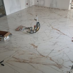 PrimeFix Homes OLEKSANDR DIACHUK - Błyszcząca, marmurowa podłoga w trakcie układania. Widoczne narzędzia i akcesoria glazurnicze: poziomice, krzyżyki dystansowe, kartony z elementami montażowymi. Fragment instalacji hydraulicznej.