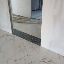 PrimeFix Homes OLEKSANDR DIACHUK - Układanie płytek podłogowych w jasnym odcieniu marmuru, z widocznymi narzędziami glazurniczymi i fragmentem szpachlowanej ściany. Widoczny fragment progu w ciemnym marmurze.