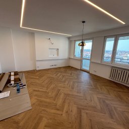 PrimeFix Homes OLEKSANDR DIACHUK - Ukończone układanie parkietu w jodełkę w jasnym pokoju z oknami i podwieszanym sufitem z oświetleniem LED. Część materiałów budowlanych widoczna w rogu.