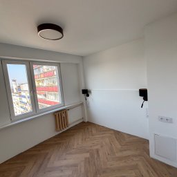 PrimeFix Homes OLEKSANDR DIACHUK - Jasne wnętrze po remoncie: parkiet ułożony w jodełkę, białe ściany z lampami ściennymi, okno z widokiem na miasto, grzejnik. Minimalistyczny design.