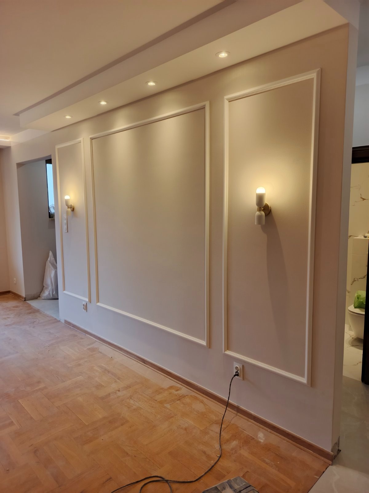 Elegancko wykończone wnętrze z panelami ściennymi, oświetleniem punktowym i drewnianą podłogą w jodełkę. Widoczne gniazdka elektryczne i fragment łazienki w tle.