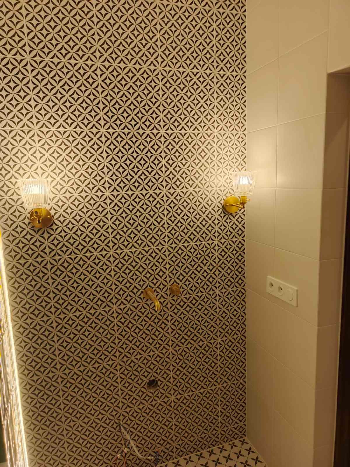 Wykończona łazienka z wzorzystymi płytkami, złotą armaturą i kinkietami. Widoczny fragment prysznica i gniazdko elektryczne. Nowoczesny design z elementami retro.