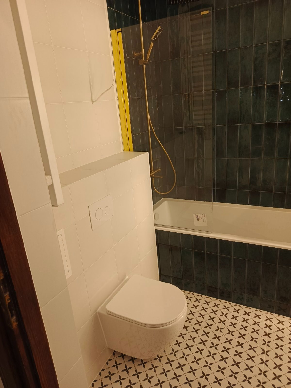 Nowoczesna łazienka z podwieszaną toaletą, ciemnozielonymi płytkami i złotą armaturą prysznicową. Wzór na podłodze dodaje charakteru wnętrzu.