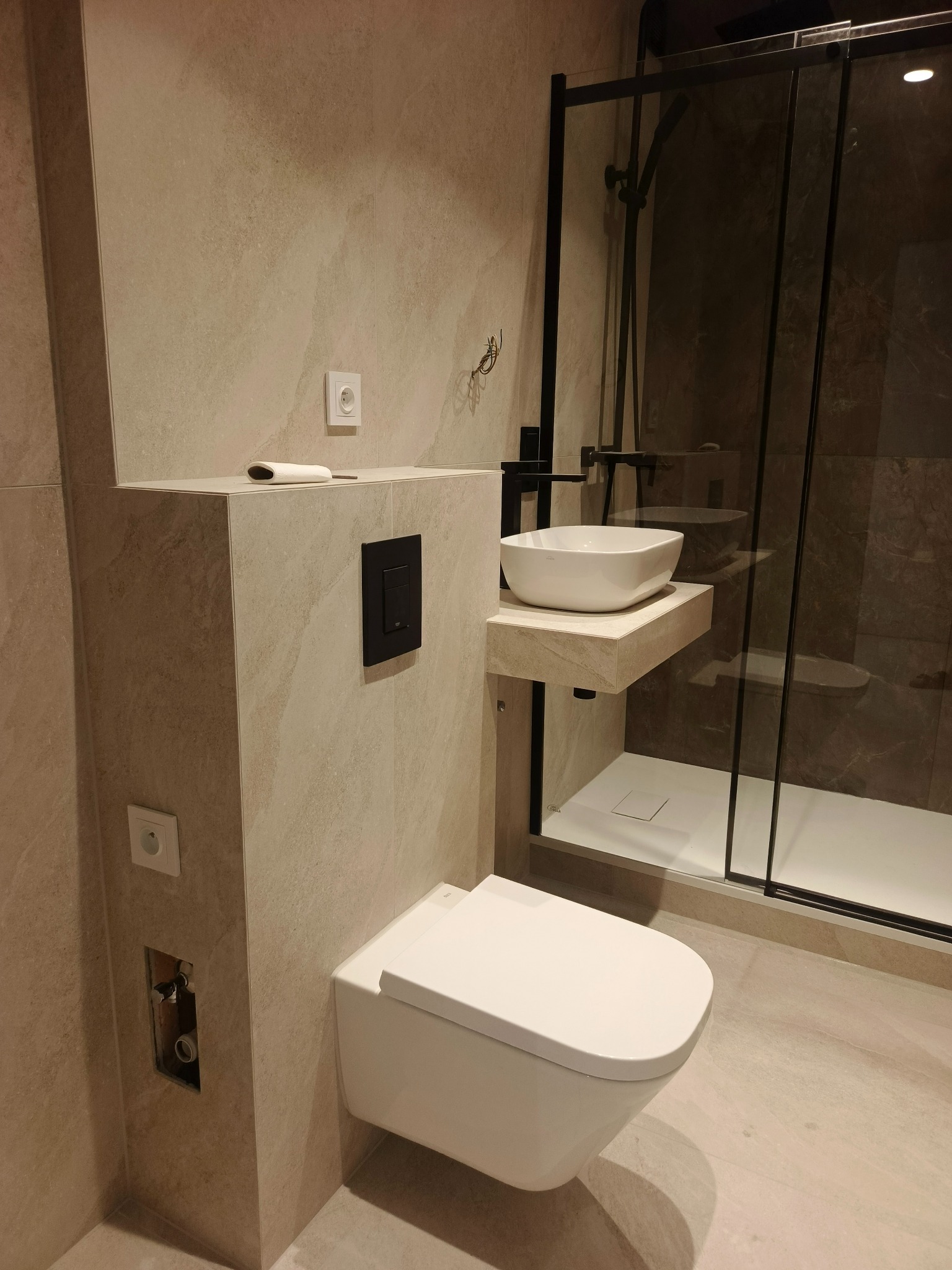 Nowoczesna łazienka w beżowych tonacjach z wiszącą toaletą, umywalką na blacie i kabiną prysznicową z czarnymi ramami. Widoczne gniazdka i czarny przycisk spłuczki.
