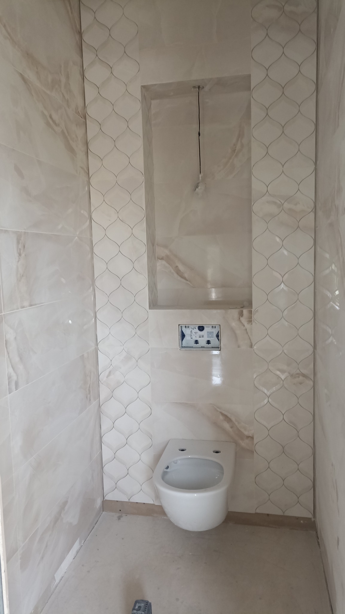 Wykończona łazienka z beżowymi płytkami imitującymi marmur, wisząca toaleta i wnęka w ścianie. Widoczne elementy instalacji podtynkowych i dekoracyjne pasy płytek.