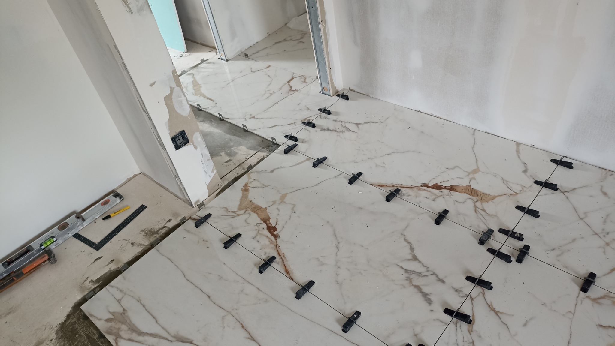 Układanie płytek podłogowych z marmurowym wzorem, z użyciem klipsów poziomujących. W tle ściany w trakcie szpachlowania i narzędzia glazurnicze.