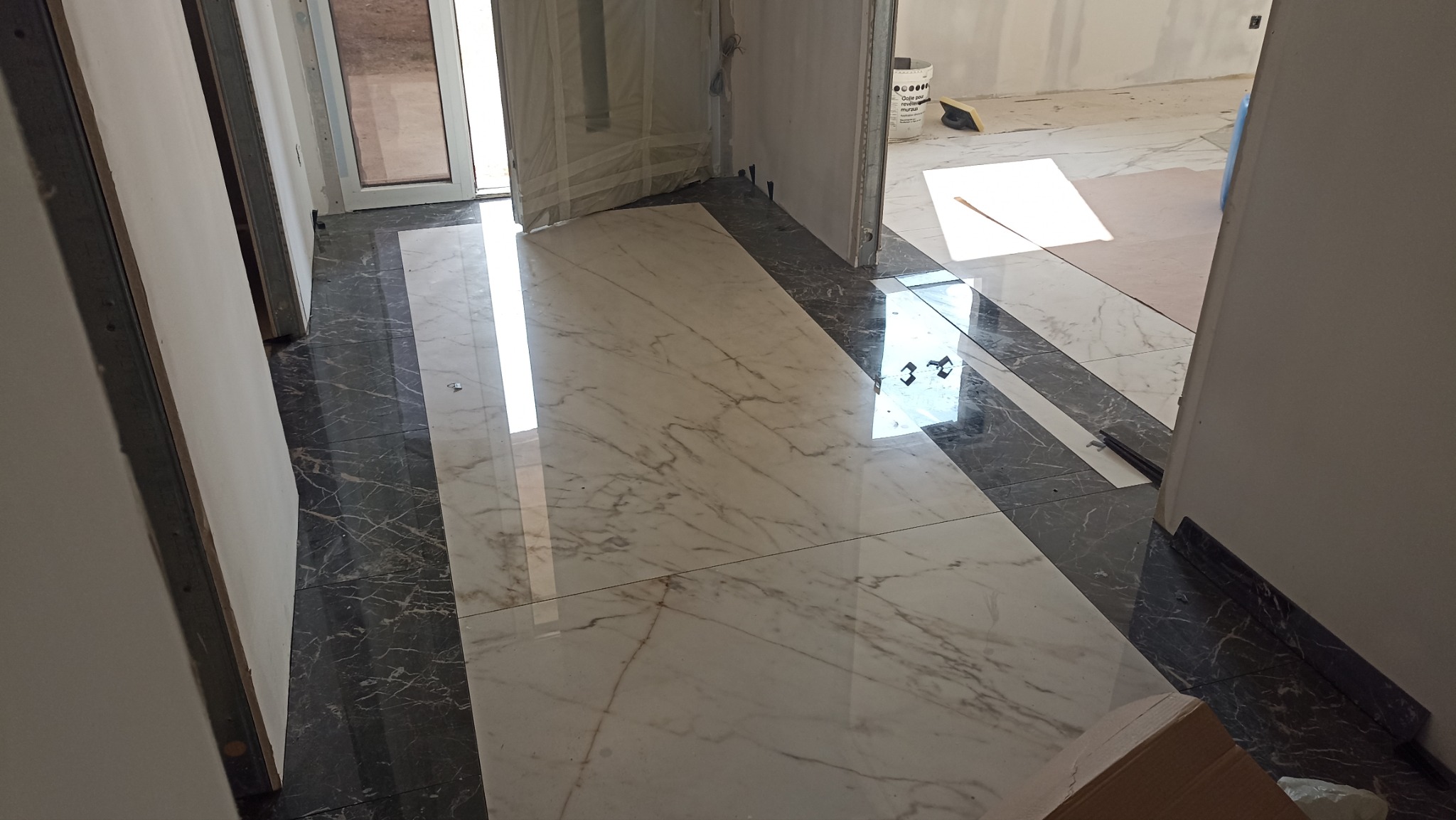 Położona, błyszcząca podłoga z marmurowych płytek w odcieniach bieli i szarości, kontrastująca z czarnymi płytkami przy ścianach. Widoczne drzwi w trakcie montażu.