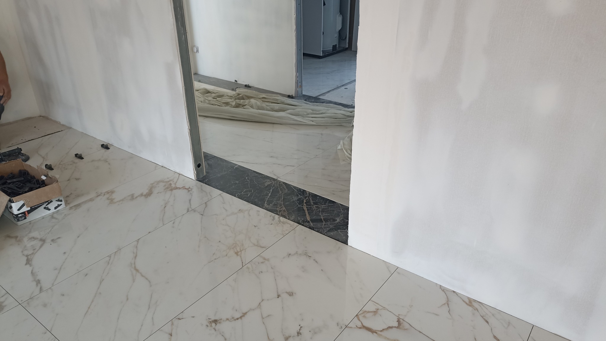 Układanie płytek podłogowych w jasnym odcieniu marmuru, z widocznymi narzędziami glazurniczymi i fragmentem szpachlowanej ściany. Widoczny fragment progu w ciemnym marmurze.