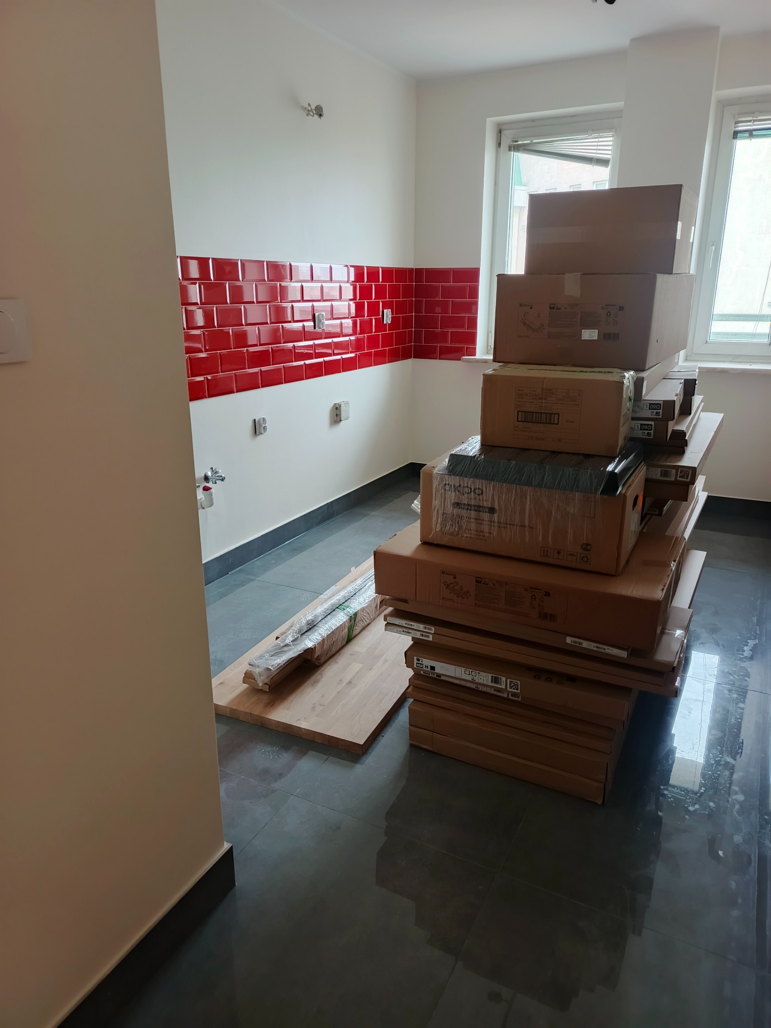 Wnętrze kuchni w trakcie remontu: czerwone płytki na ścianie, sterta kartonów z wyposażeniem na podłodze, przygotowane blaty robocze. Widok na okno.