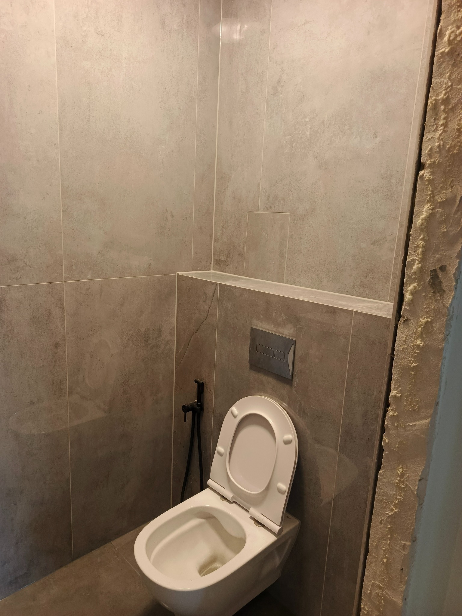 Wykończona toaleta w szarościach z bidetką, widoczna ściana w stanie surowym. Ujęcie z dołu, podkreślające nowoczesny design i etap prac wykończeniowych.