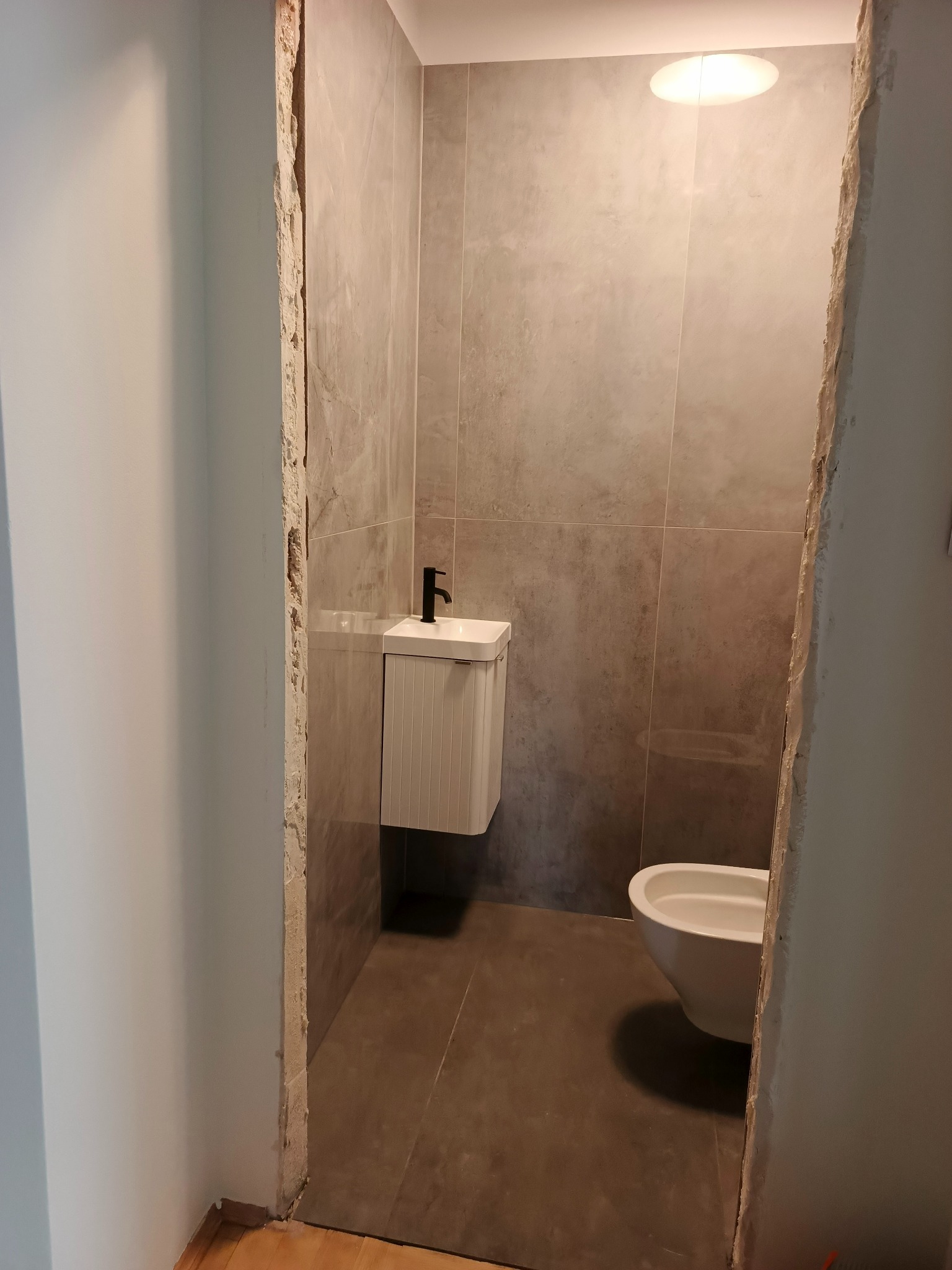 Wykończona łazienka z szarymi płytkami, wiszącą szafką z umywalką i czarną armaturą, oraz toaletą. Widoczne surowe wykończenia ościeżnicy.