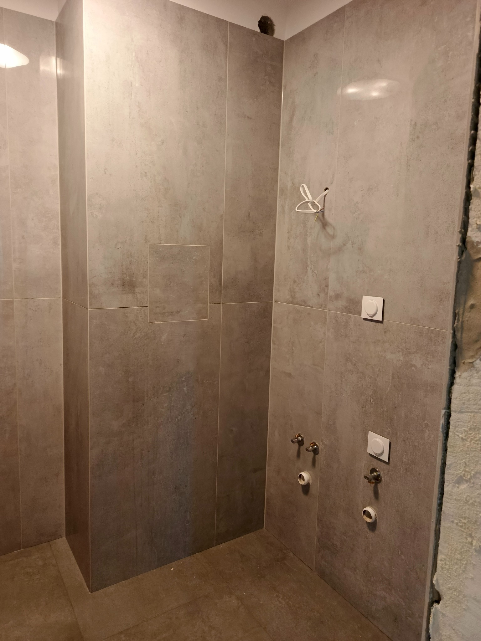 Wykończona łazienka z szarymi płytkami, widoczne przyłącza hydrauliczne i elektryczne. Ściany wyłożone płytkami imitującymi beton, surowy stan w rogu pomieszczenia.