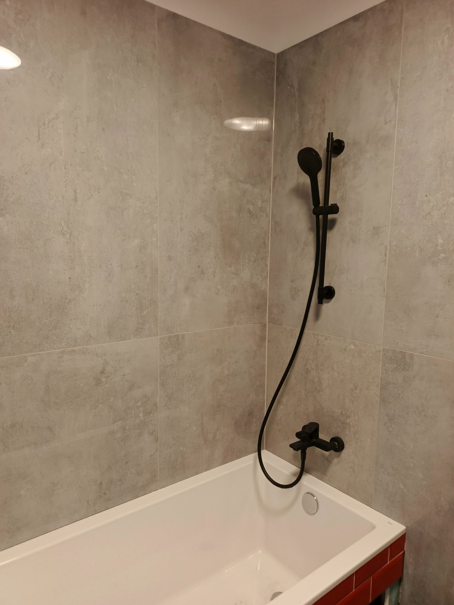 Wykończona łazienka: biała wanna z czarną armaturą, szare płytki na ścianach i czerwony akcent na obudowie wanny. Minimalistyczny design.