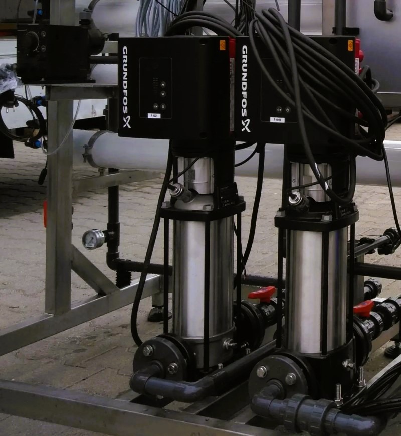 Dwie pompy Grundfos X na metalowej konstrukcji z rurami i zaworami, w tle instalacja. Widoczne przewody i elementy sterujące. Serwis pomp.