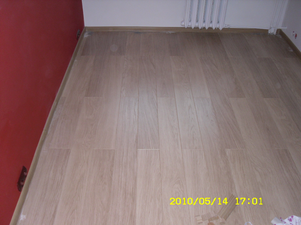 Ułożona jasna podłoga panelowa z widoczną czerwoną ścianą po lewej stronie i białym grzejnikiem u góry, z widocznymi śladami po remoncie przy ścianach.