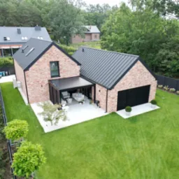 Nowoczesny dom murowany z czerwoną cegłą, czarnym dachem i garażem. Widok z góry na budynek z ogrodem, tarasem i pergolą. W tle zalesiony teren.