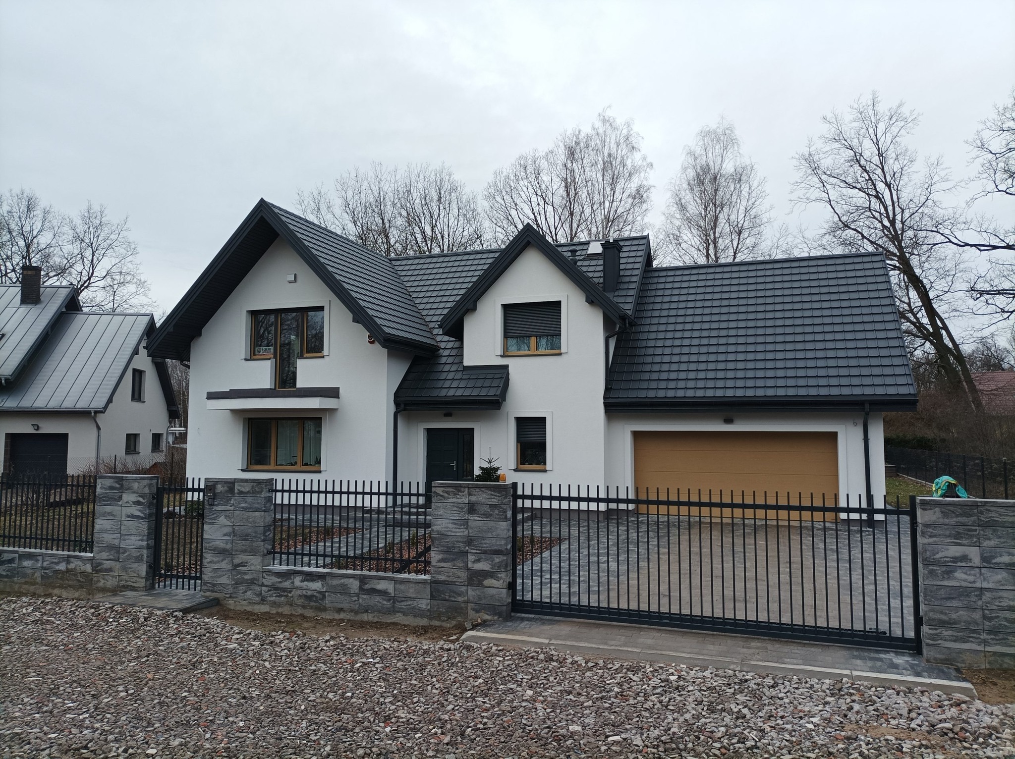 Biały dom z szarym dachem i brązową bramą garażową, otoczony czarnym ogrodzeniem z betonowymi słupkami. Podjazd wyłożony kostką, otoczenie żwirowe.