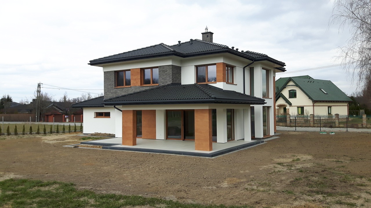 Nowoczesny dom dwukondygnacyjny z płaskim dachem, wykończony w bieli i szarości, z elementami imitującymi drewno. Widok z zewnątrz, trawnik w trakcie zakładania.