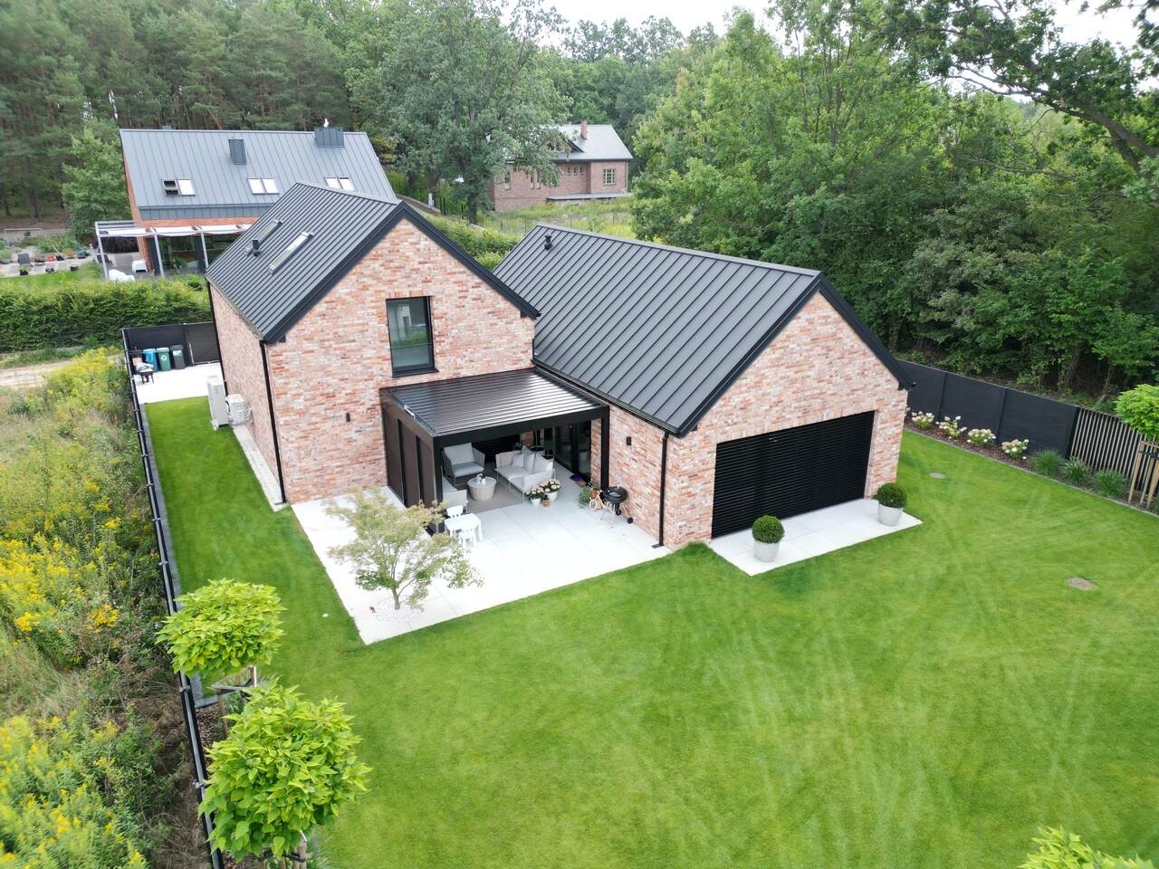 Nowoczesny dom murowany z czerwoną cegłą, czarnym dachem i garażem. Widok z góry na budynek z ogrodem, tarasem i pergolą. W tle zalesiony teren.