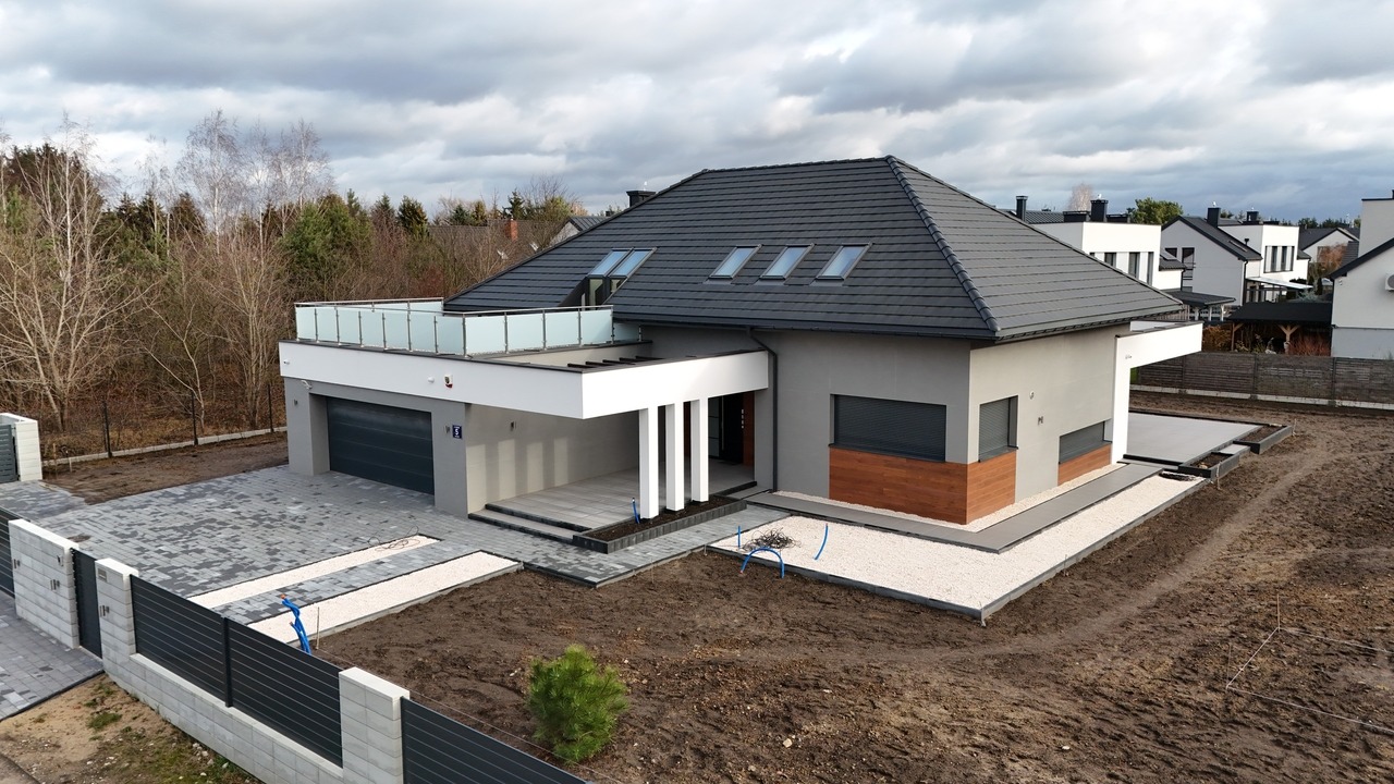 Nowoczesny, parterowy dom z płaskim dachem i tarasem na dachu. Szara elewacja, grafitowa dachówka, brama garażowa. Ułożona kostka brukowa i przygotowany teren pod ogród.