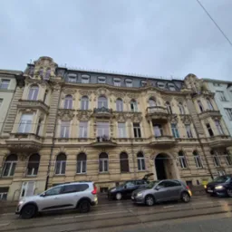 Kamienica w stylu secesyjnym z balkonami i ozdobnymi detalami architektonicznymi, widziana z perspektywy ulicy z zaparkowanymi samochodami i torami tramwajowymi.
