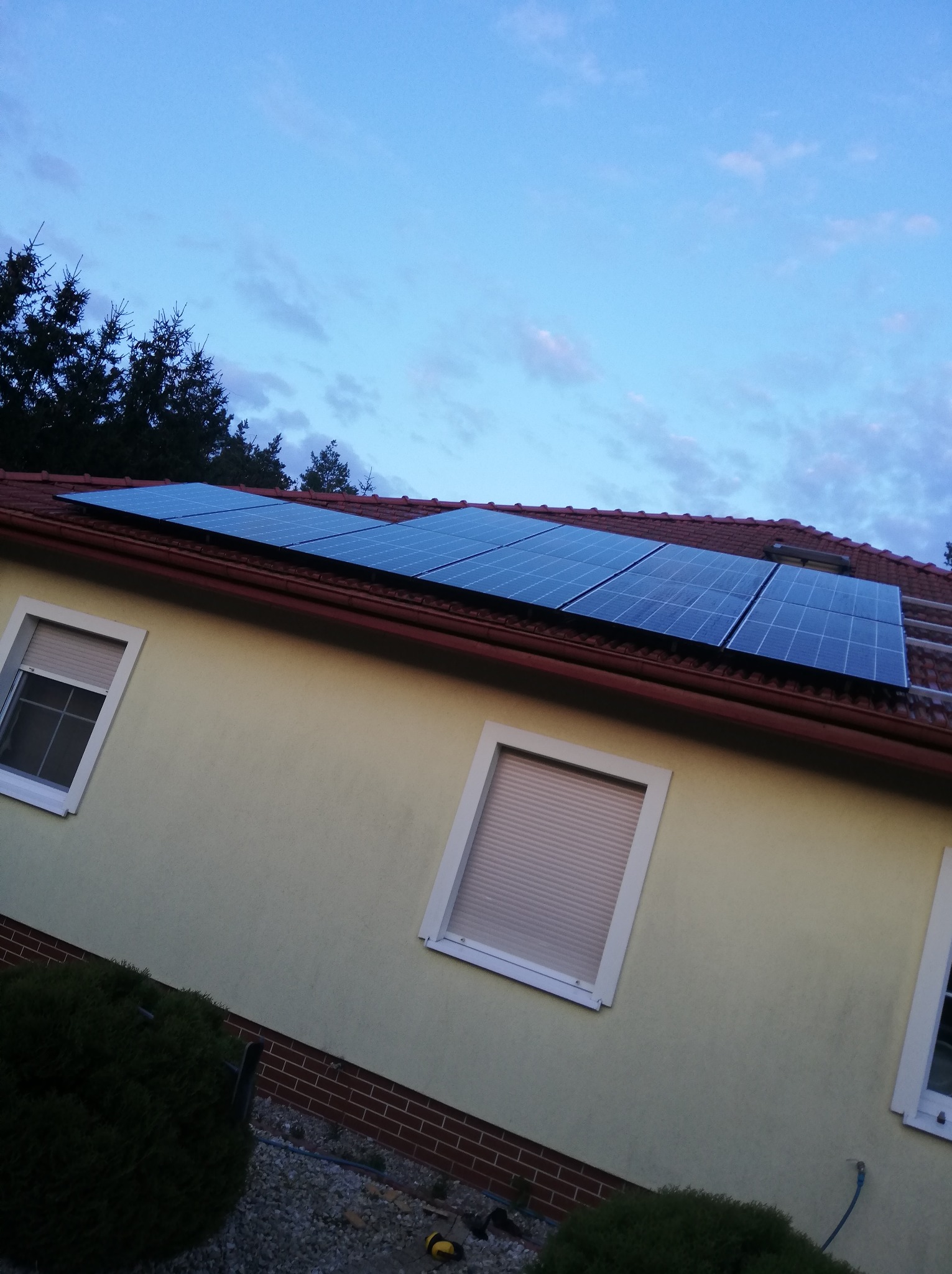 Panele słoneczne na dachu domu z czerwoną dachówką, widoczne okna z żaluzjami i fragment nieba. Ujęcie z dołu, lekko pod kątem.