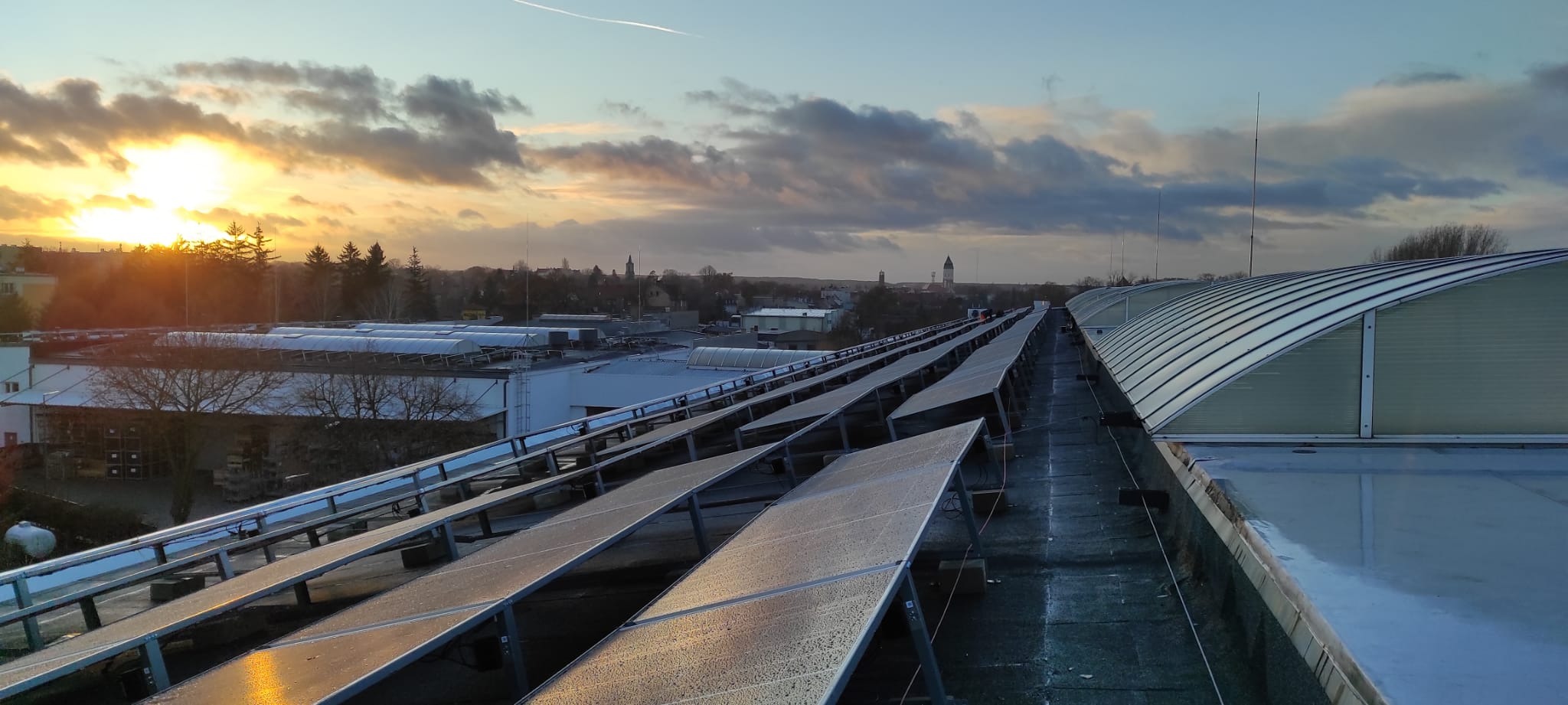 Instalacja fotowoltaiczna na dachu budynku przemysłowego o wschodzie słońca. Panele pokryte rosą. W tle widoczna zabudowa miejska i smuga kondensacyjna.