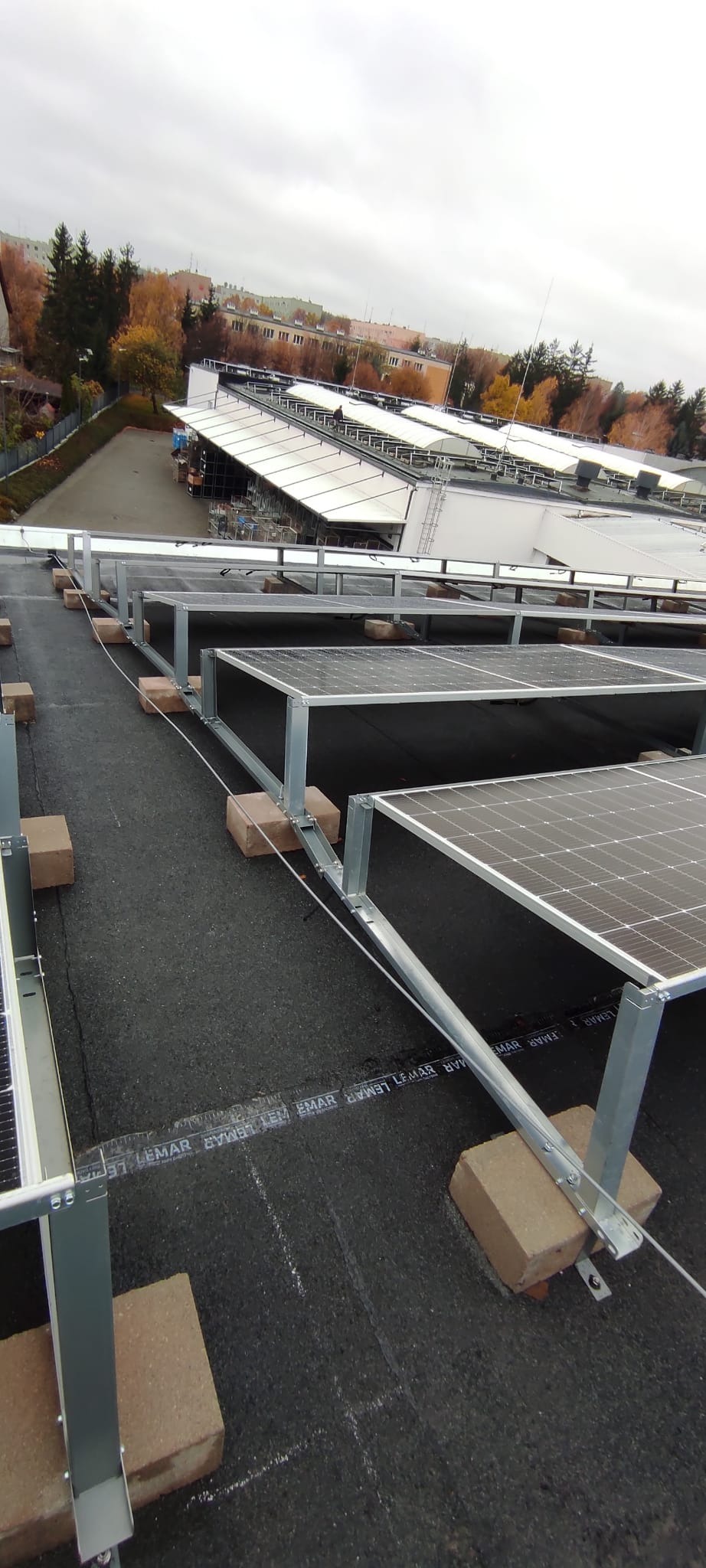Instalacja paneli słonecznych na dachu budynku z widocznymi elementami montażowymi i tłem miejskim w pochmurny dzień.