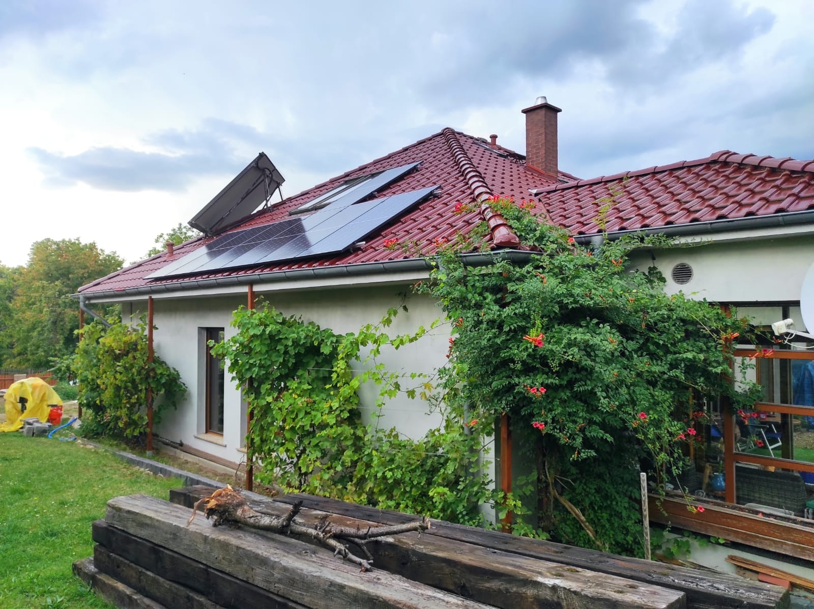 Dom z czerwoną dachówką i panelami fotowoltaicznymi na dachu, porośnięty zielenią. Na pierwszym planie stos drewna.
