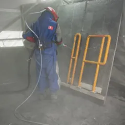 Pracownik w kombinezonie ochronnym czyści metalową konstrukcję za pomocą piaskarki. Żółta barierka zabezpieczająca obok. Widoczne efekty obróbki strumieniowo-ściernej.