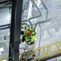 HSQ Sp. z o.o. - Pracownik w uprzęży, w kasku i kombinezonie, wykonuje prace na wysokości na tle konstrukcji przemysłowej z rurami i kratami. Ujęcie z dołu, widok na stalową konstrukcję.