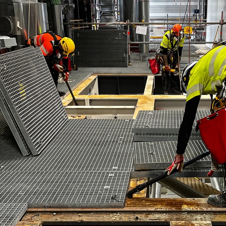 Pracownicy w kaskach i uprzężach montują metalową konstrukcję kratową na wysokości w hali przemysłowej. Widoczne narzędzia i elementy konstrukcyjne.