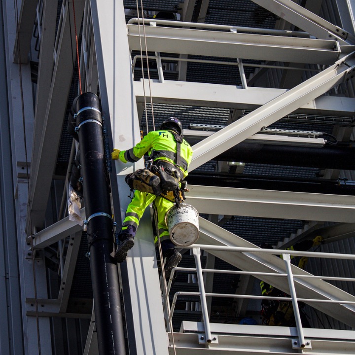 Pracownik w jaskrawożółtym kombinezonie, zabezpieczony linami, maluje konstrukcję stalową na wysokości. Widoczna puszka z farbą i elementy rusztowania.