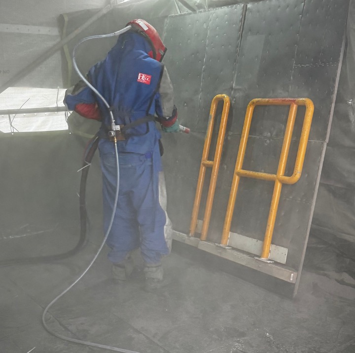 Pracownik w kombinezonie ochronnym czyści metalową konstrukcję za pomocą piaskarki. Żółta barierka zabezpieczająca obok. Widoczne efekty obróbki strumieniowo-ściernej.