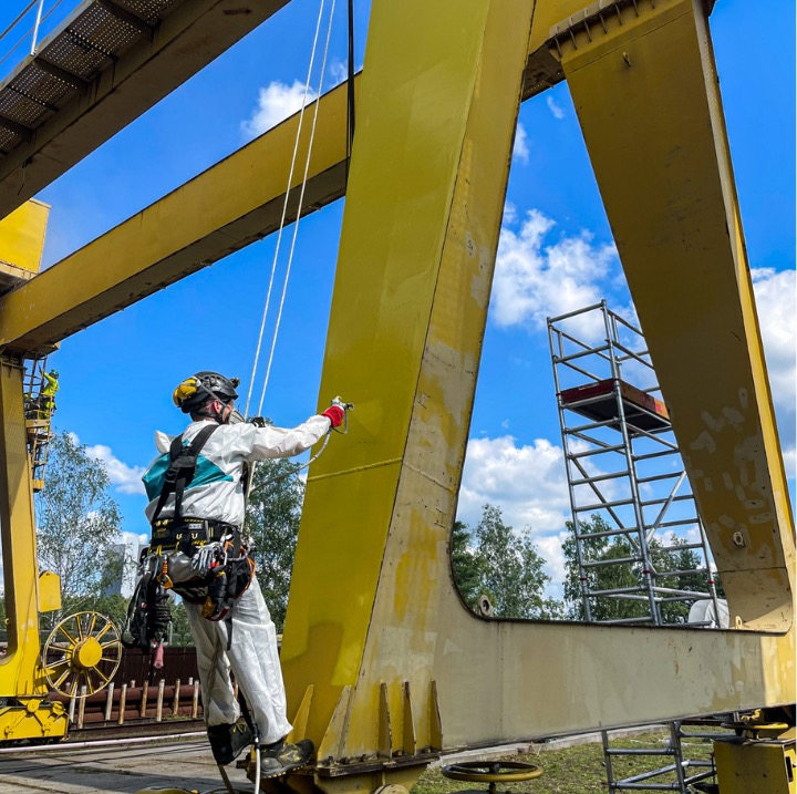 Pracownik w kombinezonie maluje konstrukcję dźwigu na wysokości, używając pistoletu natryskowego. W tle widoczne rusztowanie i błękitne niebo z chmurami.